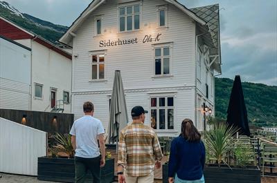 Tre personar går mot Siderhuset Ola K, ein idyllisk restaurant i Hardanger, omgitt av fjord og fjell.