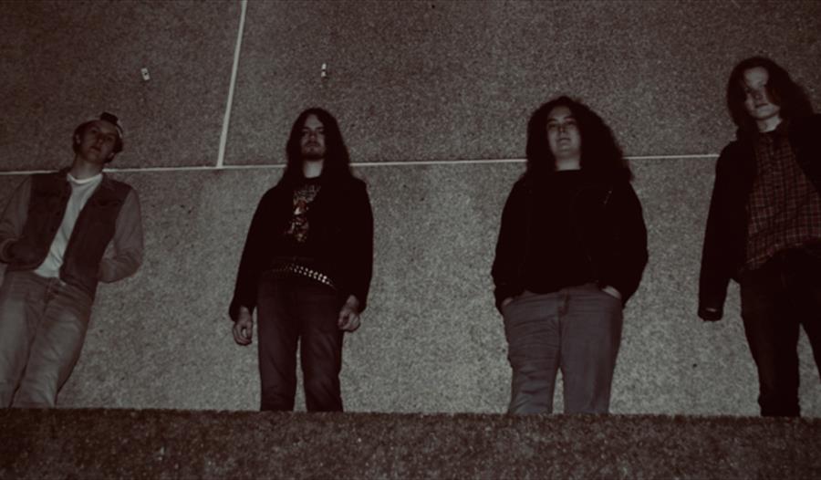 Bandmedlemmene står langs en vegg i mørket og ser ned på kameraet.