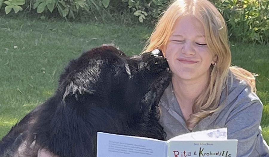 En kvinne leser en barnebok og klemmer en stor svart hund