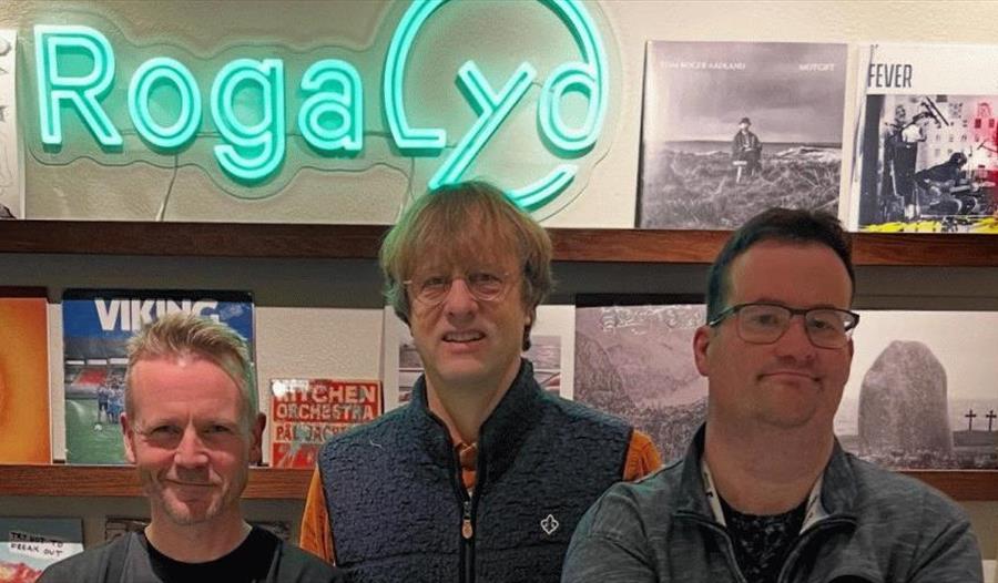 Tre menn som står foran hyllen med vinylplater. Rogalyd er på toppen av bildet