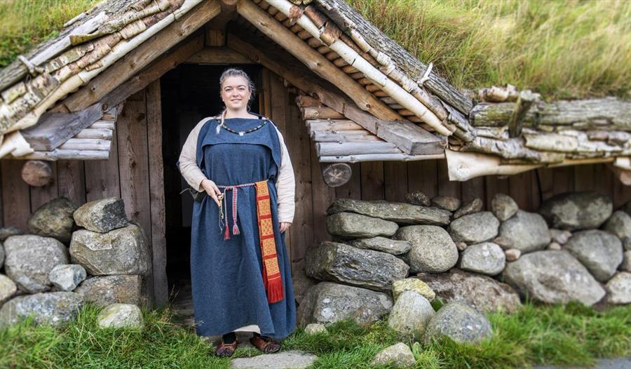 En kvinne i vikingdrakt foran huset på Jernaldergårdens huset