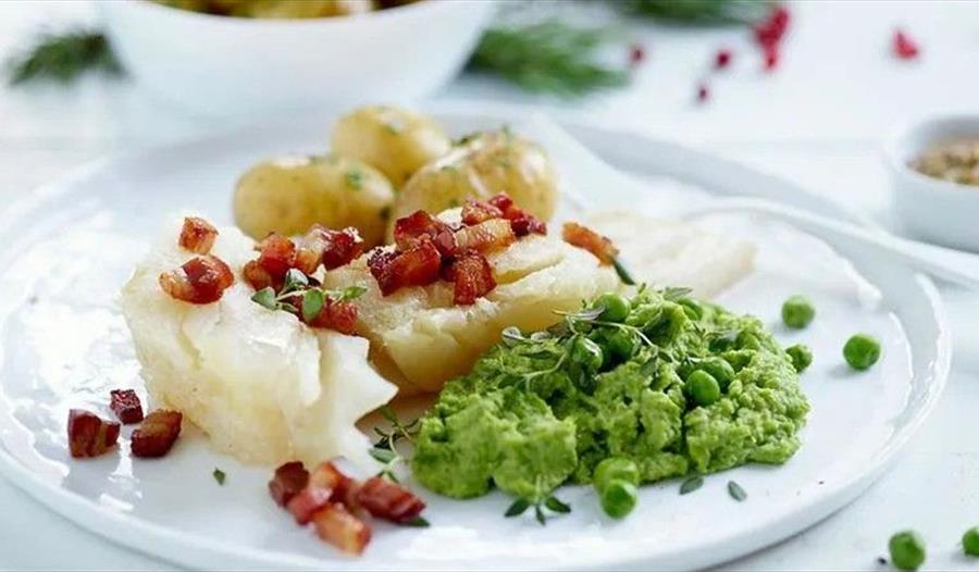 En hvit tallerken med lutefisk, bacon strødd over, ertestuing, og poteter.