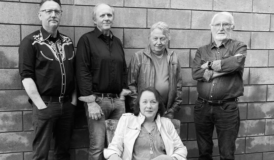Sølvi Bakken sitter i lenestolen, mens Remember band står på ryggen