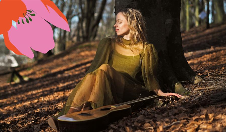 Stina Kjelstad sitter lent mot et tre, og det ligger en gitar ved siden av henne