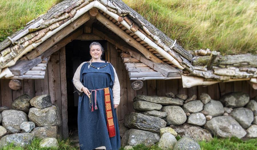 En kvinne i vikingdrakt foran huset på Jernaldergårdens huset