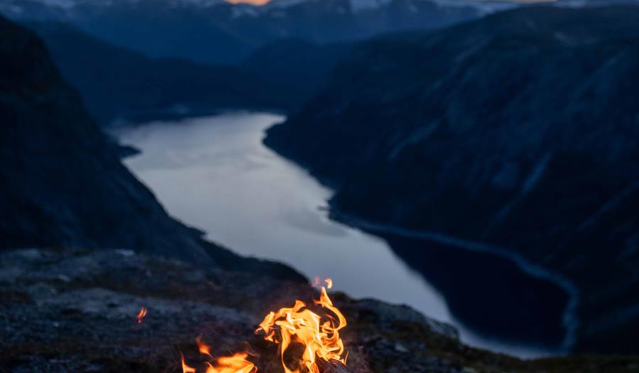 Bålpanne med åpen ild på en fjelltopp ved solnedgang med utsikt over en fjord.