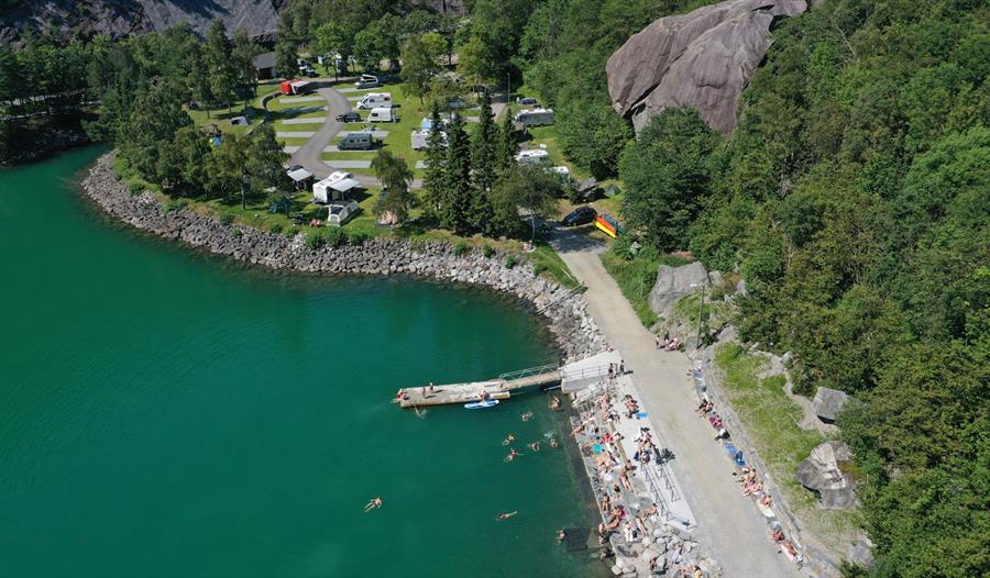 Badeplass ved Trolltunga Camping med brygge, badande gjester og campingplassen langs vatnet.
