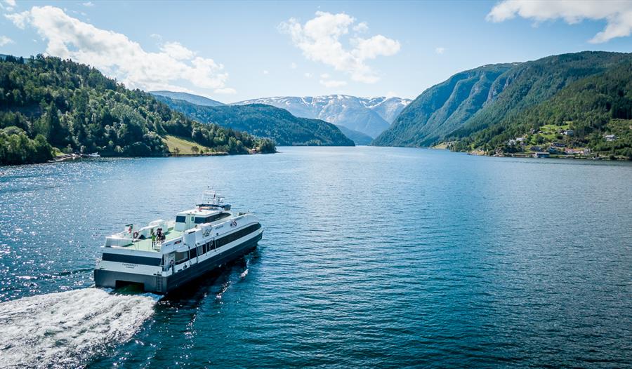 Fjordcruisebåten Fjorddrott seiler innover Hardangerfjorden med snødekte fjell i bakgrunnen, sett frå drone.