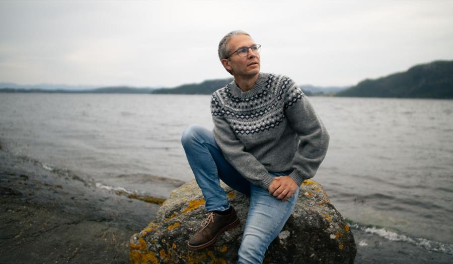 En ung mann med grå strikkegenser, jeans og briller som sitter på en stein i fjære med havet som bakgrunn.
