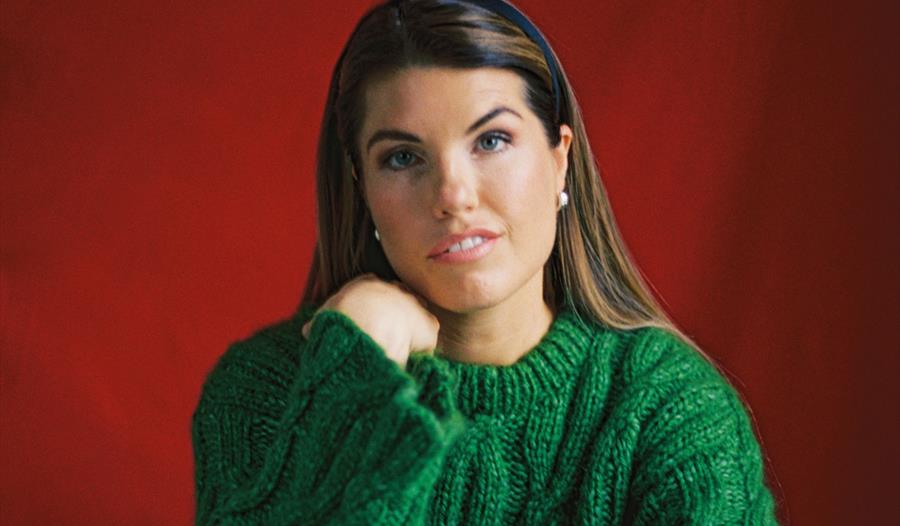 En dame med brunt hår kledd i en grønn genser foran en rød bakgrunn