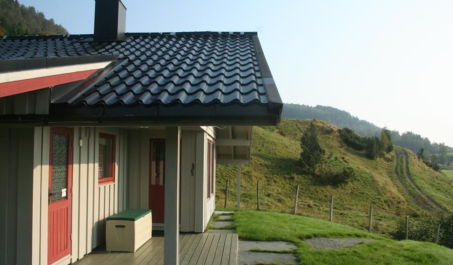 Tradisjonell norsk hytte med overbygd terrasse, raude dører og utsikt mot grøne åsar i Hardanger.