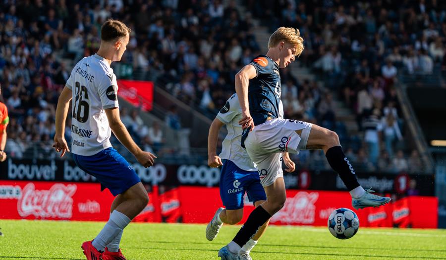 En fotballspiller spiller ballen mellom to motspillere foran fullsatte tribuner