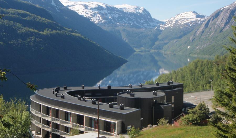 Hordatun Hotel sett frå sommarlandskap, med utsikt mot Røldalsvatnet og dei snødekte fjelltoppane i bakgrunnen.