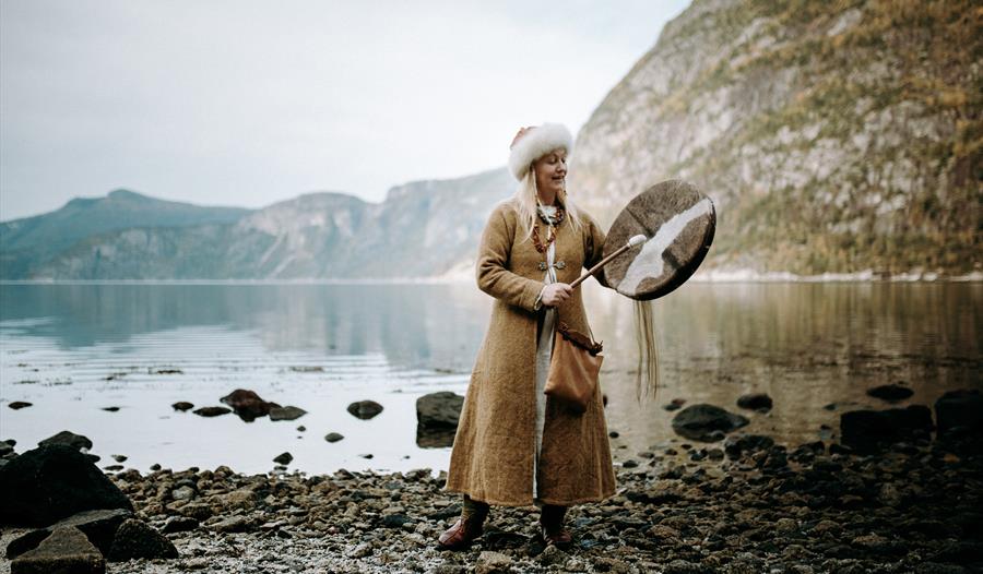 En kvinne utkledd som en viking, som står ved fjorden og spiller en vikingtromme.