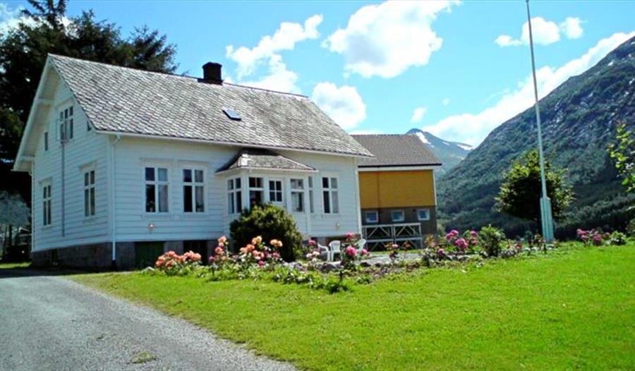 Tradisjonell kvit bygning på Dønhaug Gjestegard, omgjeve av grøn plen og med blå himmel i bakgrunnen. Perfekt for ein roleg ferie i Hardanger.