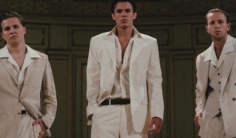 Tre menn kledd i hvite dress på en scene, bandet Bausa