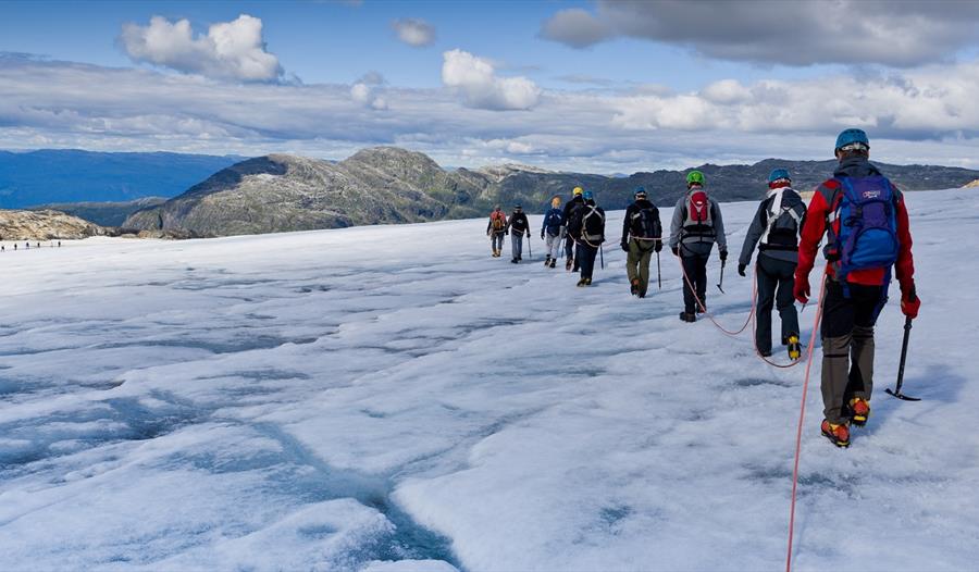 Gruppe med fotturistar på tur over Folgefonna-breen i Hardanger, leia av ein breførar med isøks og stegjern.