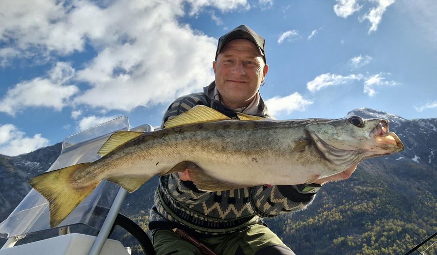 En mann smiler mens han holder en stor fisk ombord på en båt med fjell i bakgrunnen.
