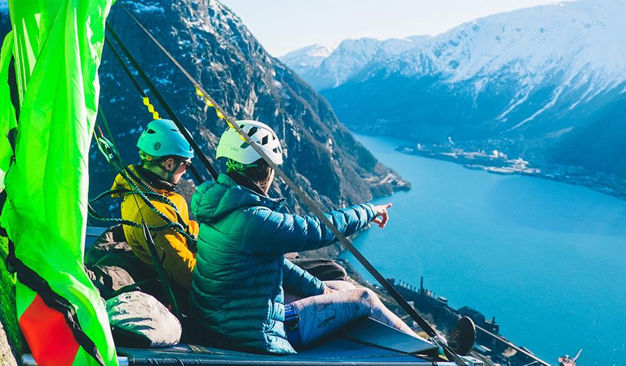 To personar sit på ein portaledge ved fjorden i Hardanger, med majestetiske fjell i bakgrunnen. Ein unik overnattingoppleving i fjellveggen.