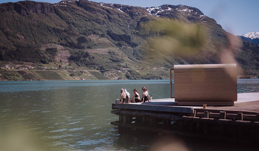 Personer nyter fjordbad på brygge foran Heit Grimo sauna med fjell i bakgrunnen