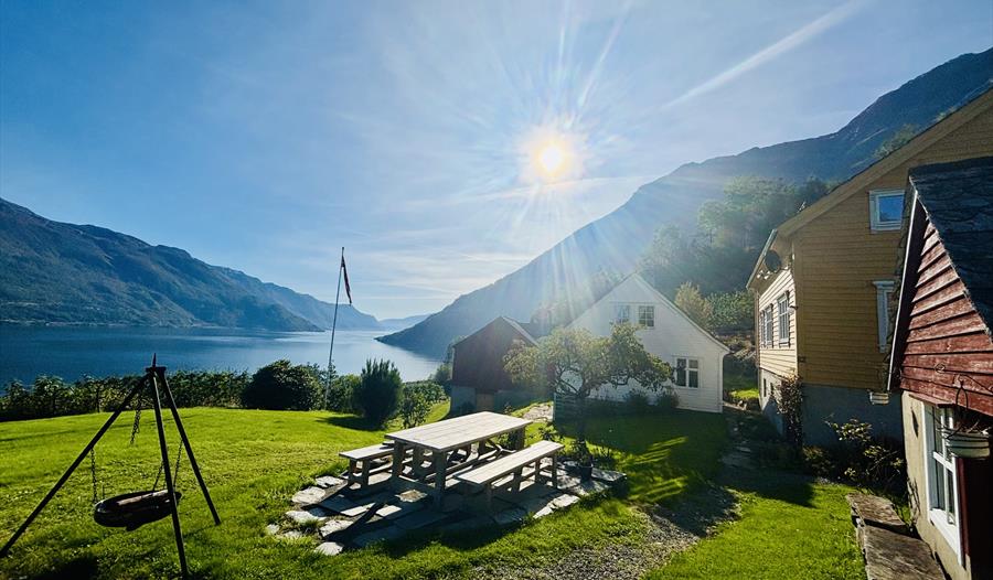 Solfylt uteområde ved Jaastad Gjestetun med sitjeplassar, grønt tun og utsikt over Hardangerfjorden og fjella.