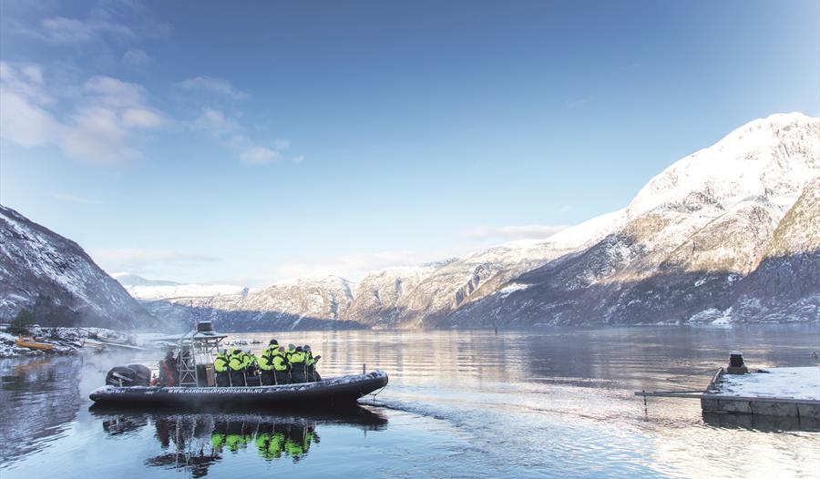 Et vinterbilde av en ribbåt fylt med passasjerer, snøfylte fjell i bakgrunnen.