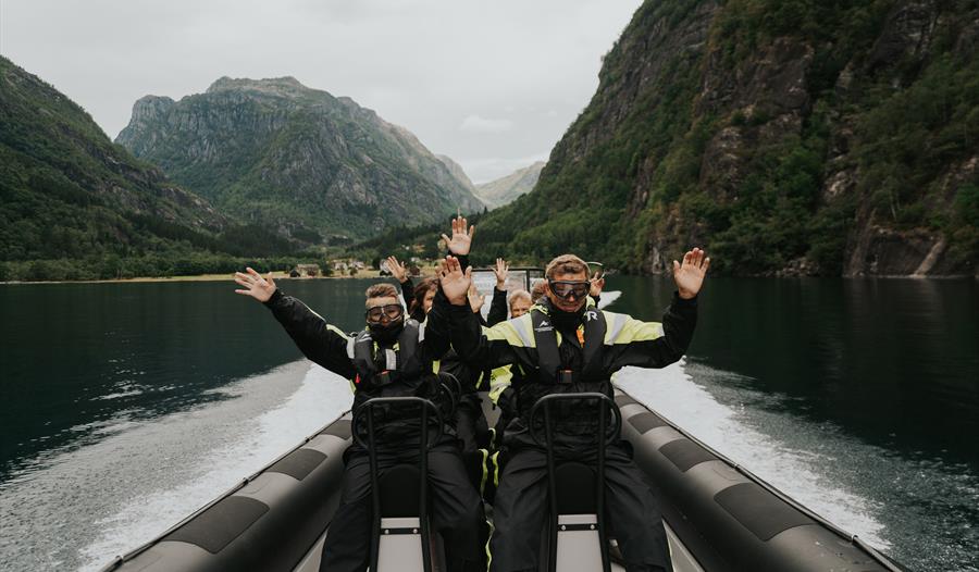 Glade turistar løftar armane i glede medan dei køyrer gjennom fjorden i Hardanger.