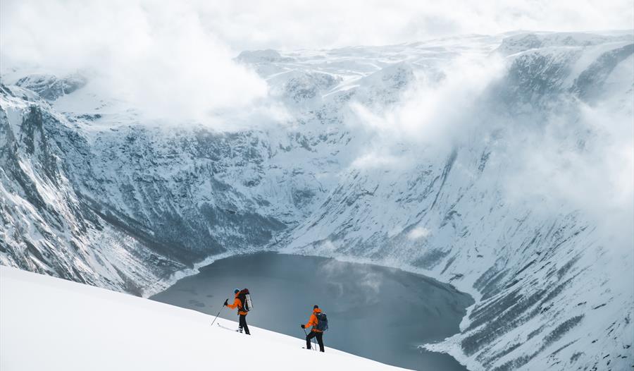 To skigåere ved et vann omringet av snøkledde fjell.