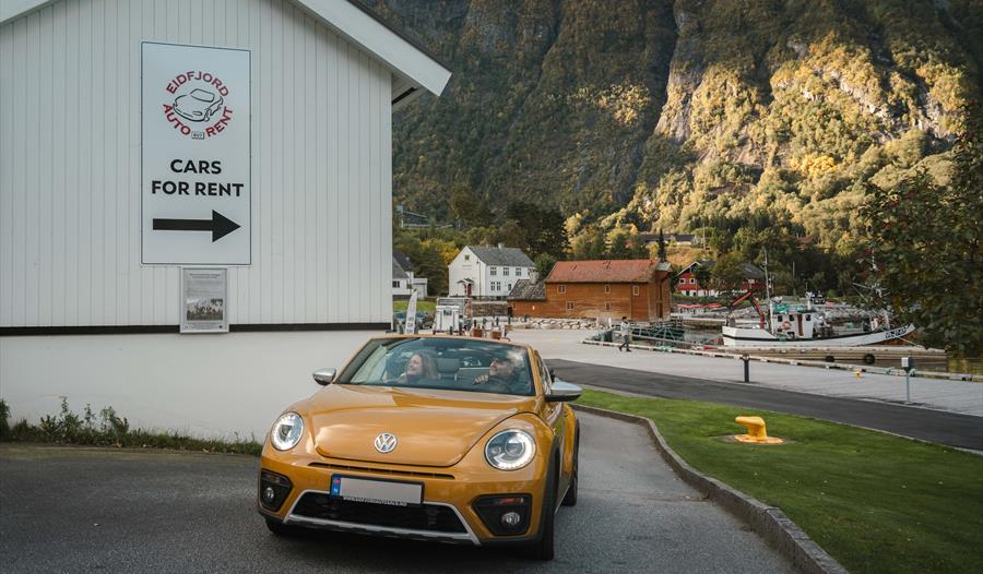 Et par i en gullfarget VW Beetle-bil under et skilt hvor det står «Biler til leie».
