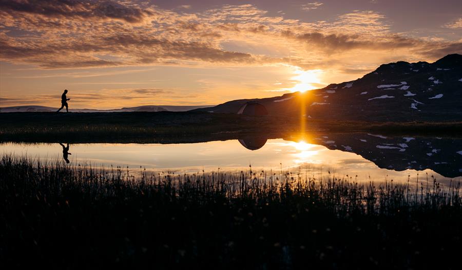 Menneske går ved kanten av et vann inn mot solnedgangen.