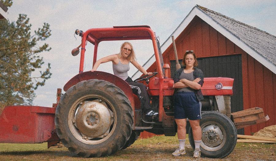 Mari Strand Ferstad står sitter i en traktor foran en låve, mens Malin Landa står ved siden av traktoren.