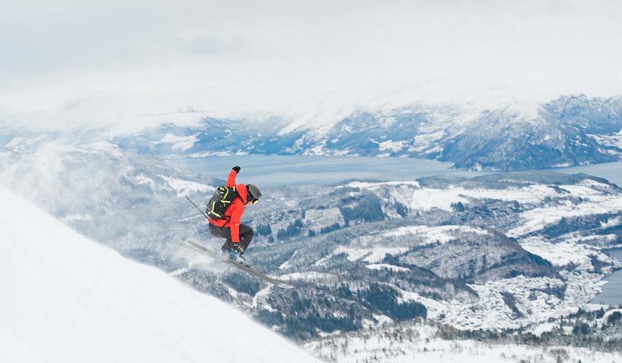 Topptur i Hardanger. Skikjører hopper ned en fjellside med utsikt mot Hardangerfjorden og snødekket landskap.