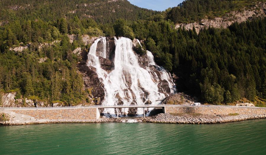 Furubergsfossen frå fjorden – Eit storslått bilete av Furubergsfossen som renn nedover klippene ved fjorden i Kvinnherad, omgitt av frodig natur.