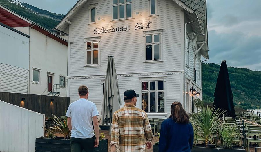 Tre personar går mot Siderhuset Ola K, ein idyllisk restaurant i Hardanger, omgitt av fjord og fjell.