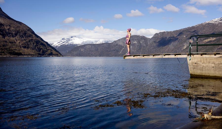 En kvinne kledd i lilla vinterbikini, hansker og lue, stående på stupebrettet ved Kråkeskarvet strand.