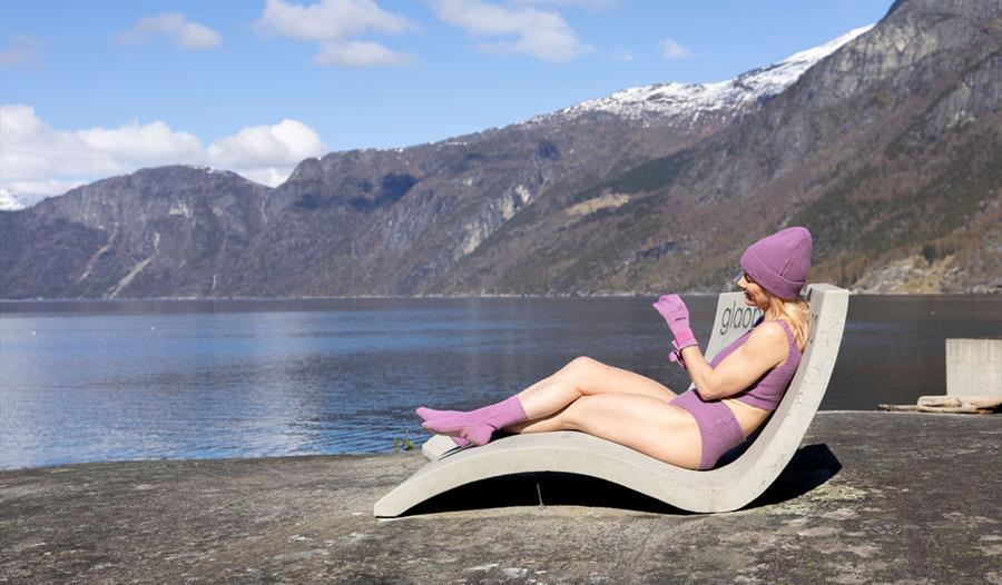 En kvinne kledd i lilla vinterbikini, hansker og lue, sittende i en av betongstolene på Kråkeskarvet strand.