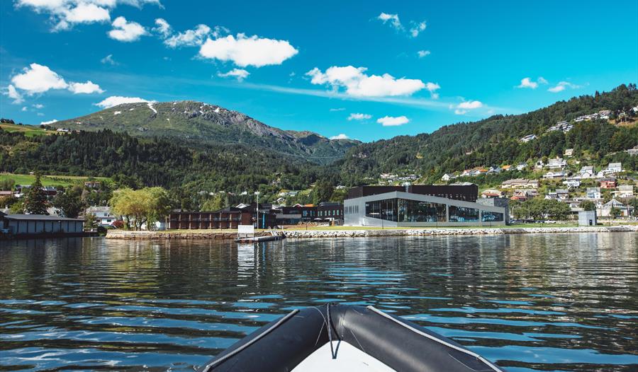 Utsikt mot Hardangerfjord Hotel frå fjorden, med fjell og bygd i bakgrunnen.