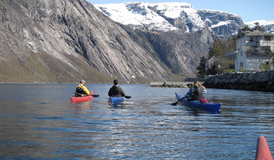 Kajakkpadlere i Eidfjord med snødekte fjell i bakgrunnen og et tradisjonelt norsk hus ved siden av fjorden.