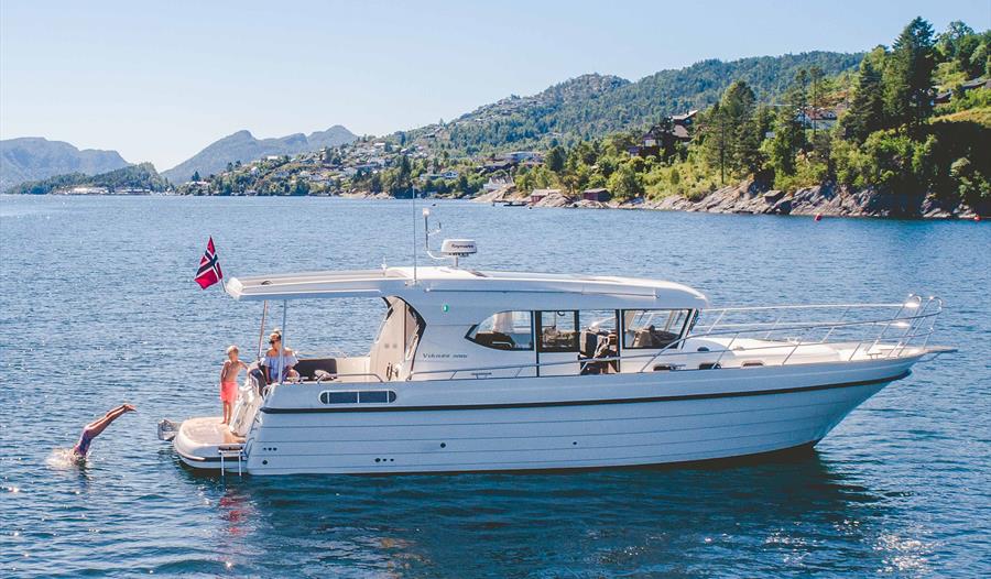 Båten Pilagutt i Hardangerfjorden med sol, blått vatn og folk som badar. Perfekt for ein avslappande fjordcruiseoppleving.
