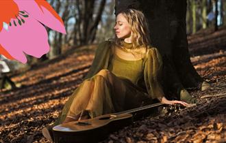 Stina Kjelstad sitter lent mot et tre, og det ligger en gitar ved siden av henne