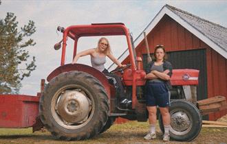 Mari Strand Ferstad står sitter i en traktor foran en låve, mens Malin Landa står ved siden av traktoren.