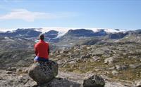 En rødkledd mann sitter på en stein og ser mot Hardangerjøkulen isbre.