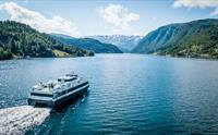 Fjordcruisebåten Fjorddrott seiler innover Hardangerfjorden med snødekte fjell i bakgrunnen, sett frå drone.