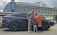En familie står foran en bil. I bakgrunnen står det et cruiseskip.
