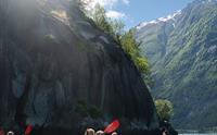 Gruppe mennesker padler i gule kajakker langs en bratt fjellvegg under blå himmel.