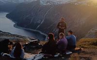 En gruppe mennesker sitter på fjellet og ser på solnedgangen over en fjord.