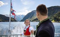 Par i bunad held kvarandre i handa på akterdekket under fjordcruise på Hardangerfjorden, norsk flagg i bakgrunnen.
