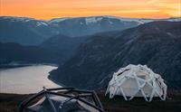 Geodetiske telt på fjelltoppen ved solnedgang med utsikt over en fjord.