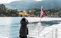 Passasjer fotograferer fjordlandskapet frå akterdekket under fjordcruise på Hardangerfjorden, norsk flagg i vinden.