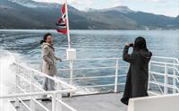 To passasjerar tar selfie på akterdekket under fjordcruise på Hardangerfjorden, snødekte fjell i bakgrunnen.